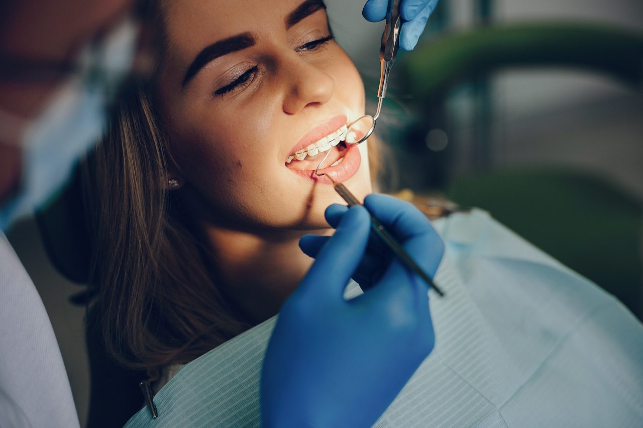 Moça jovem está no dentista, ela usa aparelho metálico com as borrachinhas transparente, o dnetista checa com espelhinho.