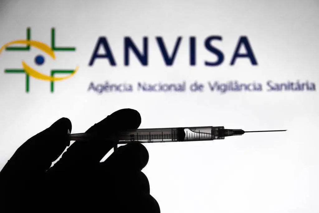 Pessoa de luva apertando seringa. Ao fundo a logo da ANVISA.