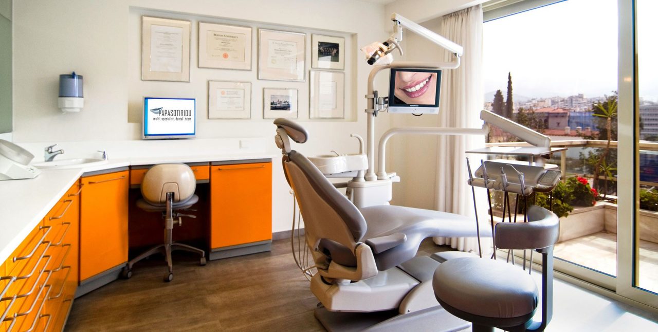 Consultório odontológico com vista para cidade, tem uma sacada. No consultório tem escrivaninha com computador, várias divisórias no armário, certificados na parede, a cadeira odontológica com equipo no centro.