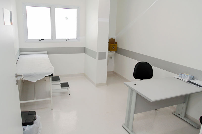 Ambulatório de um hospital. A sala é toda branca, tem maca e uma cadeira com mesa para o médico.