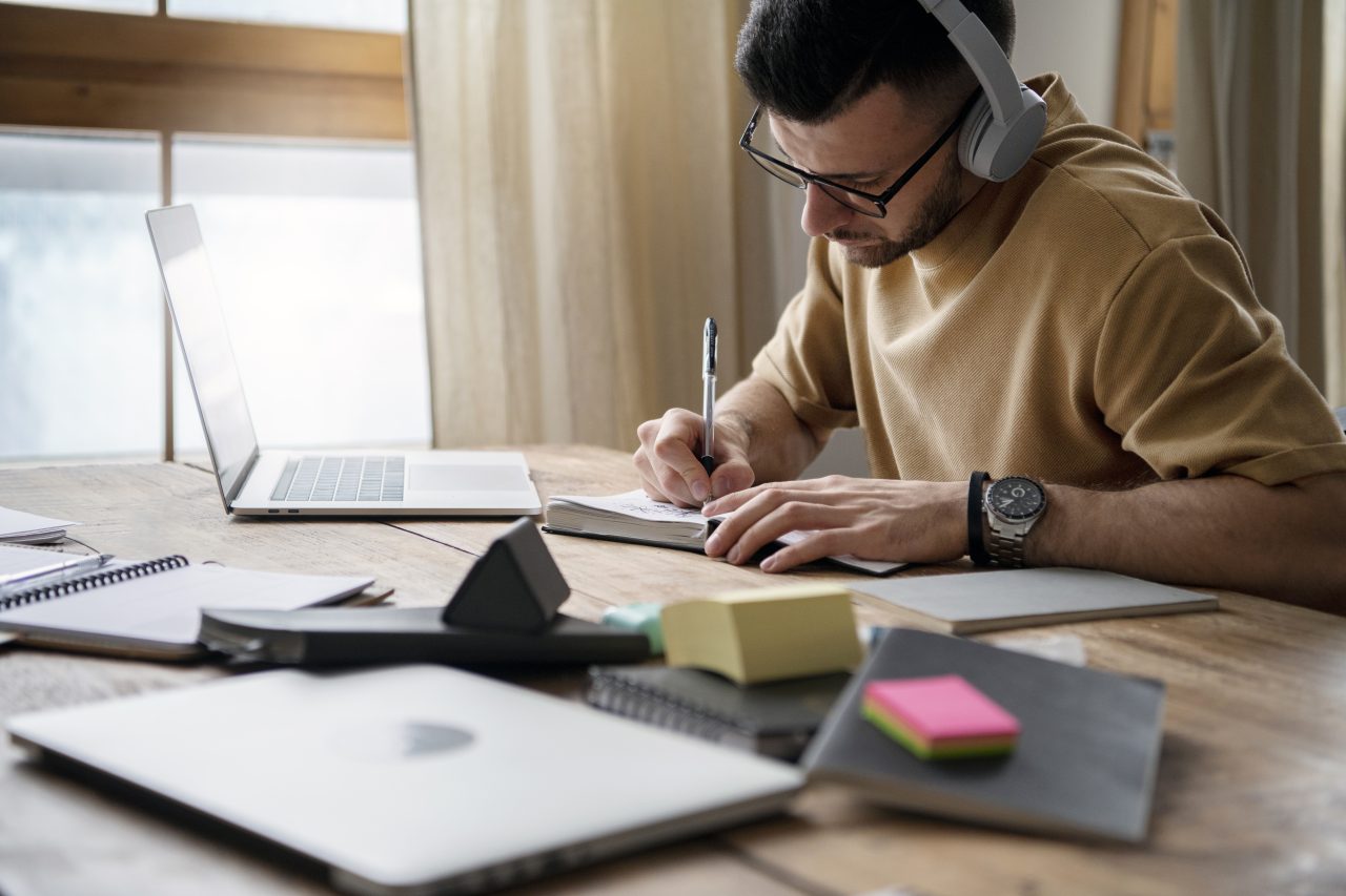 Homem sentado em frente a mesa de madeira com vários livros, cadernos, post-its. Ele está usando um fone de ouvido e na frente dele tem um caderno no qual ele está escrevendo com caneta e ao lado um notebook aberto.