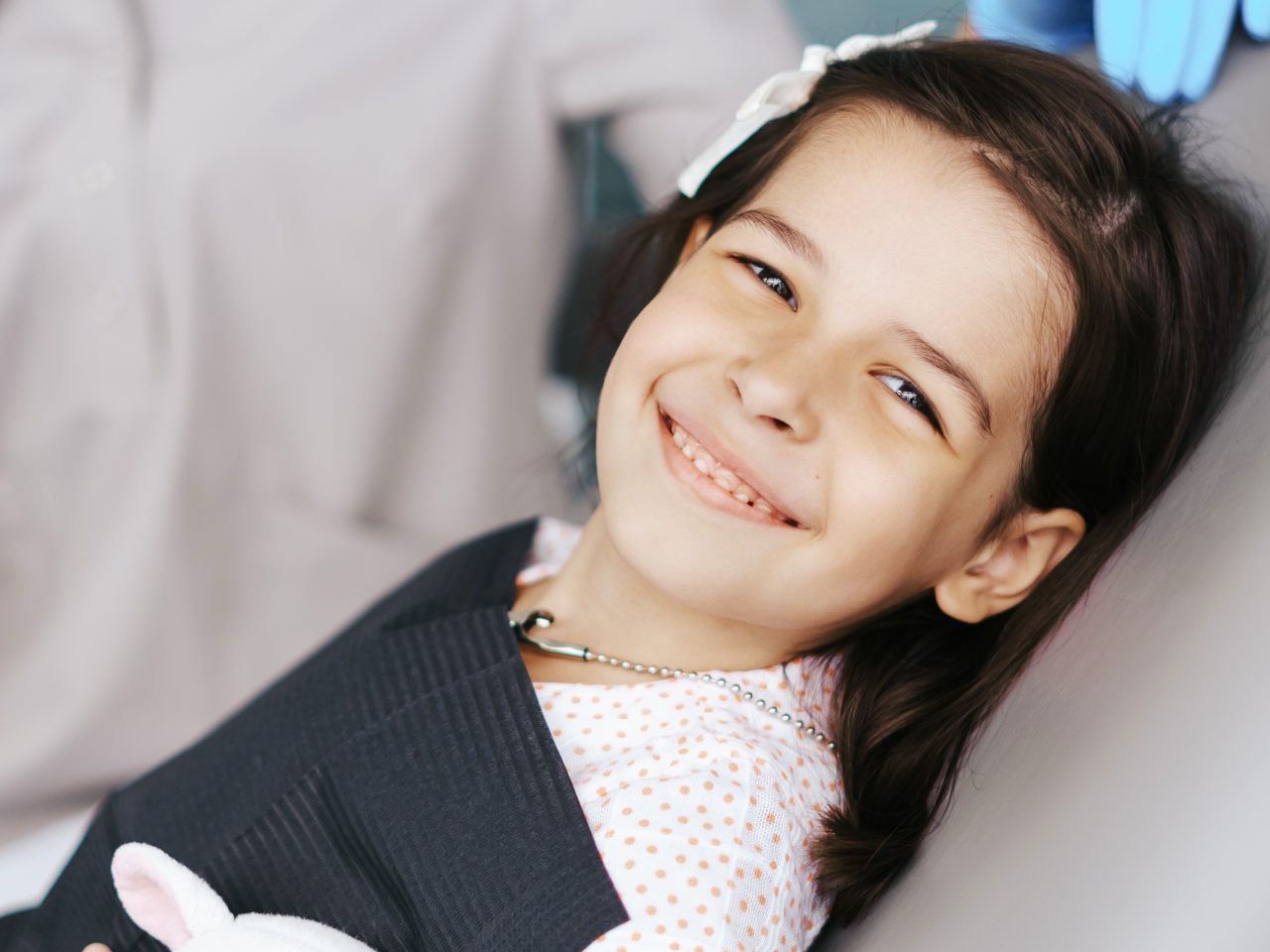 Menina está na cadeira odontológica segurando um coelhinho de pelúcia e sorrindo.