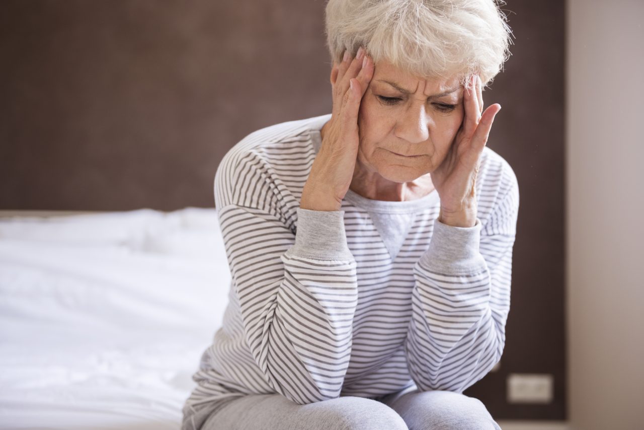 Senhora idosa sentada na beira da cama com os cotovelos apoiados no joelho e a mão na cabeça expressando dor de cabeça.