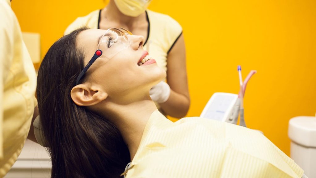 mulher deitada para fazer procedimento em dentista, de fundo pode se ver dentista mulher, a sala é toda amarela