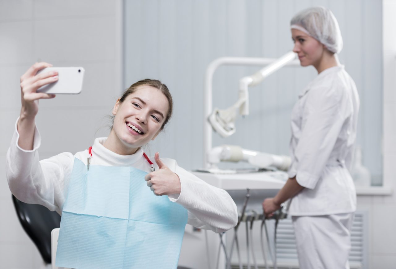 dentista no fundo e paciente sentada na cadeira sorrindo e tirando selfie