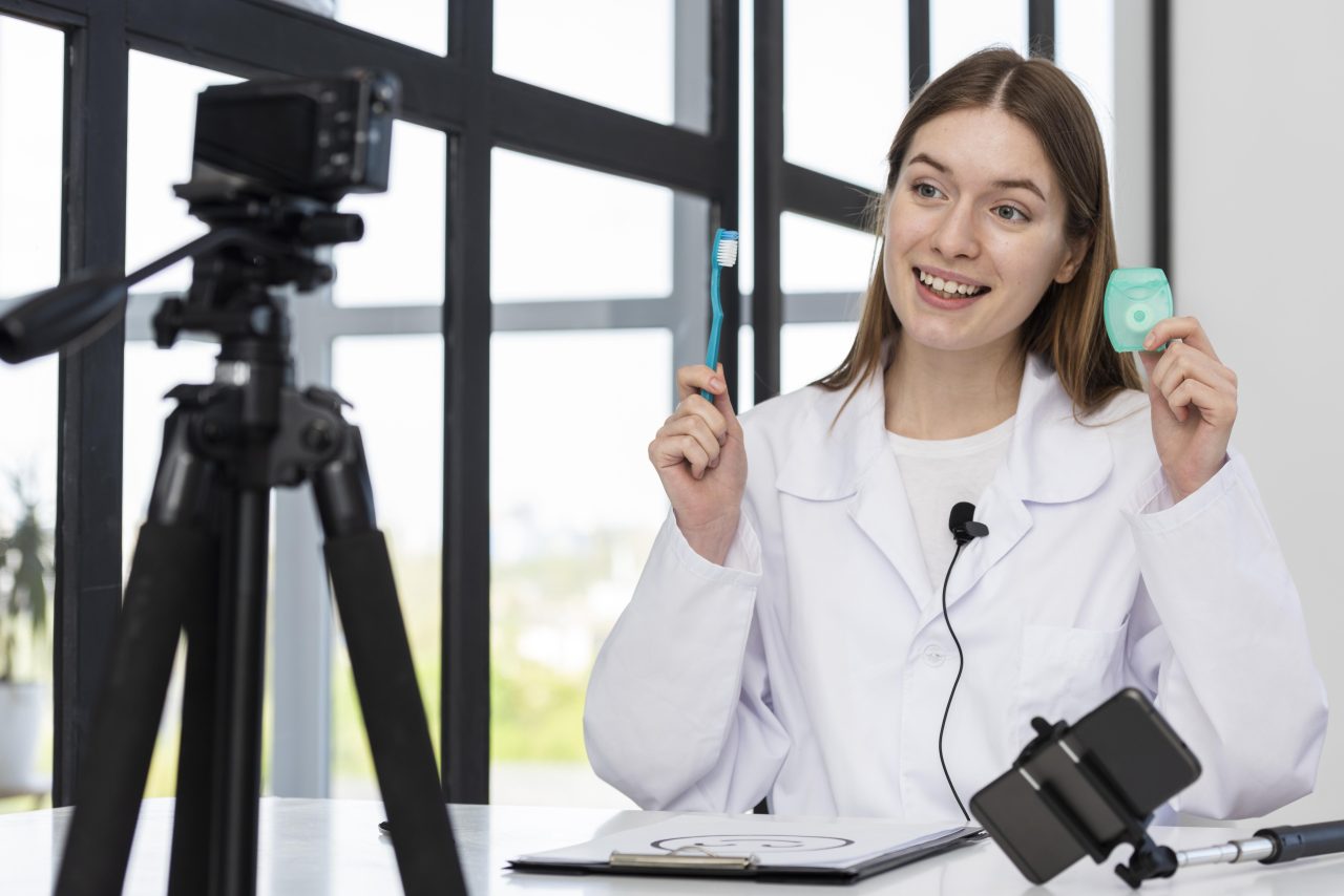 Dentista mulher está segurando escova de dente e fio dental, está com um microfone de lapela e sentada gravando vídeo de reels para dentistas.