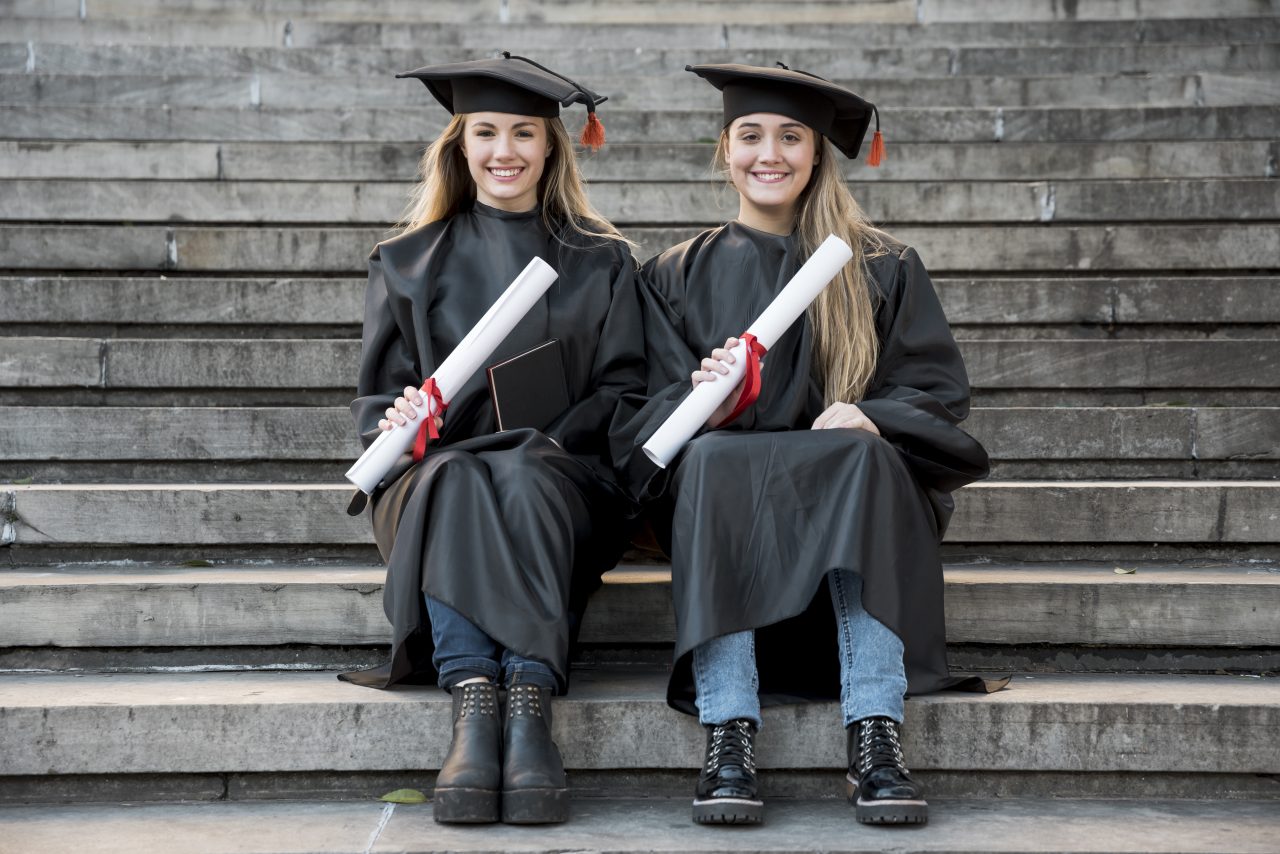 Duas mulheres loiras sentadas em uma escadaria usando beca de formatura e segurando seus diplomas.