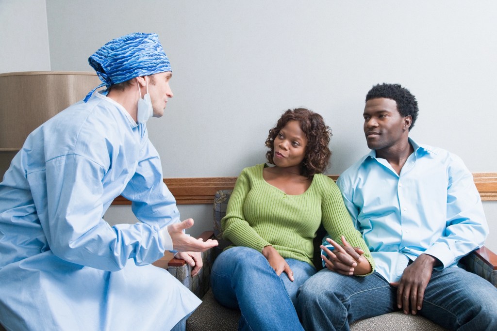 Doutor usando roupa de cirurgia e touca azul está sentado na sala de espera conversando com casal de pacientes, os quais estão de mãos dadas.