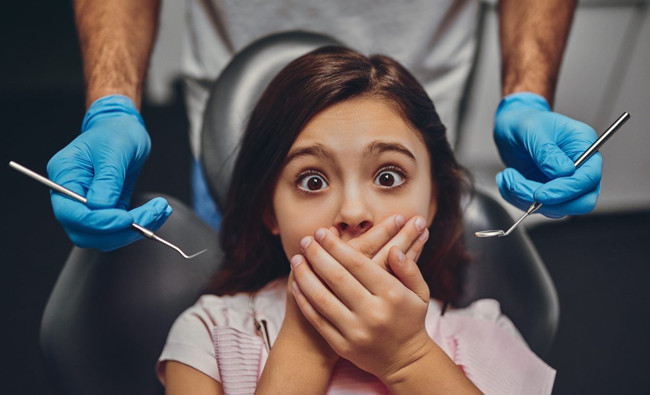 Garotinha com olhos arregalados e cobrindo a boca com as duas mãos. Ela está no dentista com medo. Atrás dela tem um dentista.