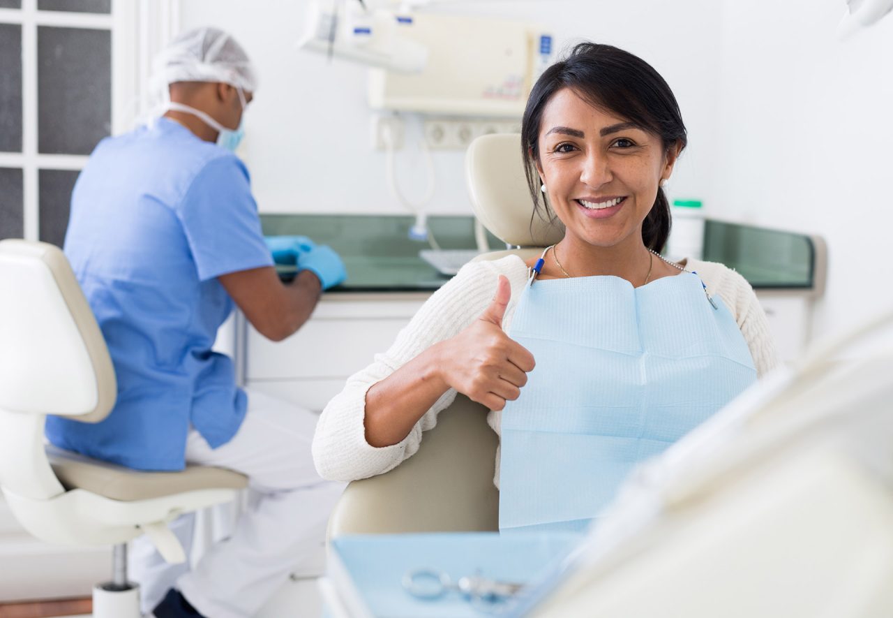 Paciente é uma mulher de suéter branco, cabelos lisos castanhos escuros e está fazendo sinal de positivo com o dedão. No fundo, o dentista está arrumando algo.