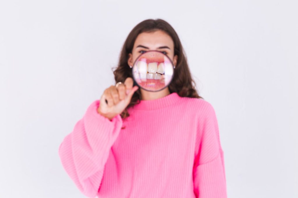 mulher de suéter rosa segurando lupa e, frente e a boca que dá aumento em seus dentes