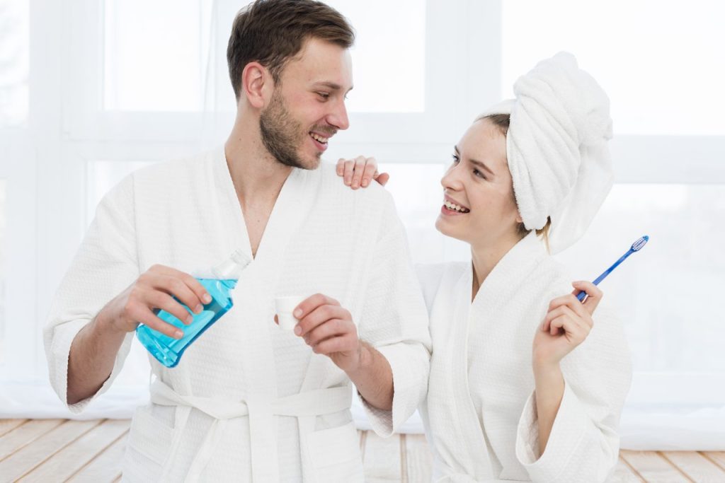 homem e mulher no banheiro usando robes, ela está segurando uma escova de dente e ele um enxaguante bucal