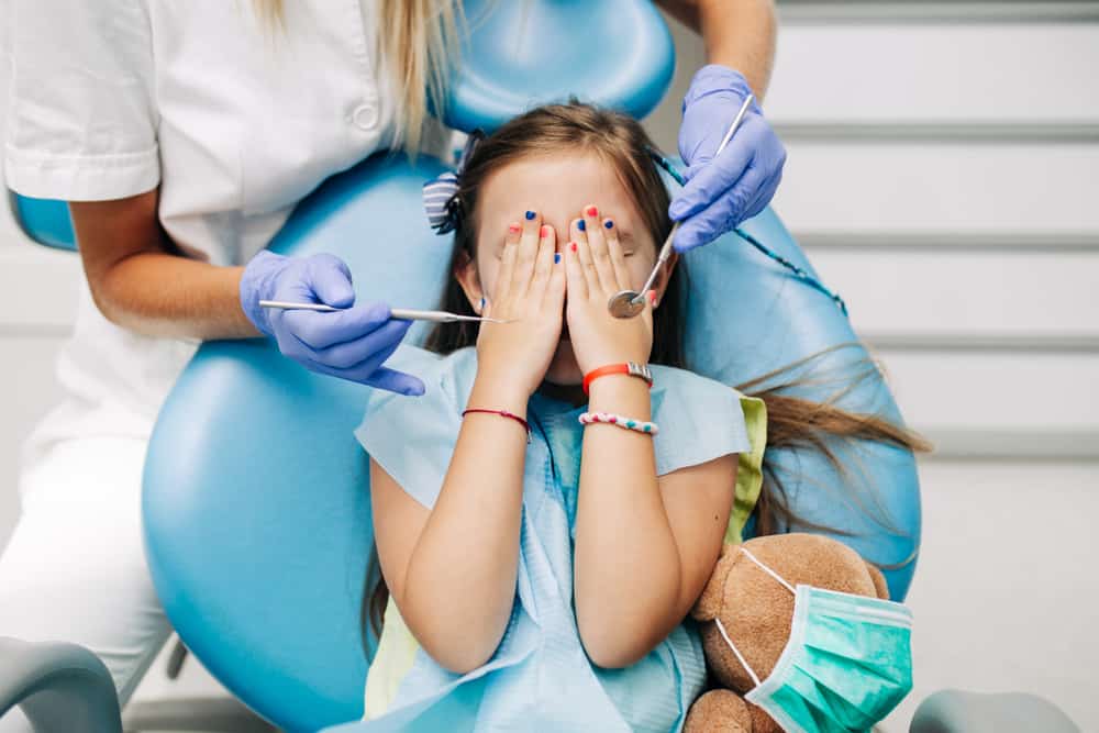 Garotinha loira cobre o rosto com as mãos, com medo da dentista.