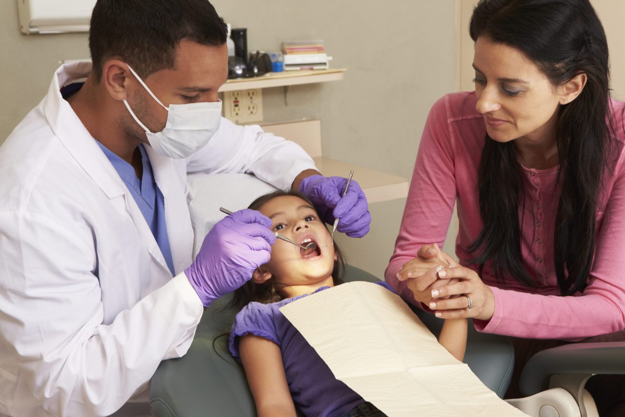 Mãe segurando mão da filha criança durante procedimento odontológico.