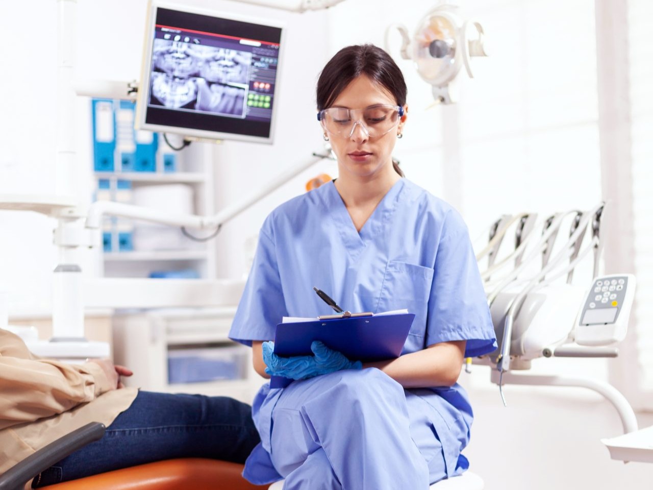 Dentista sentada anotando em sua prancheta, enquanto a paciente está deitada na cadeira do consultório.
