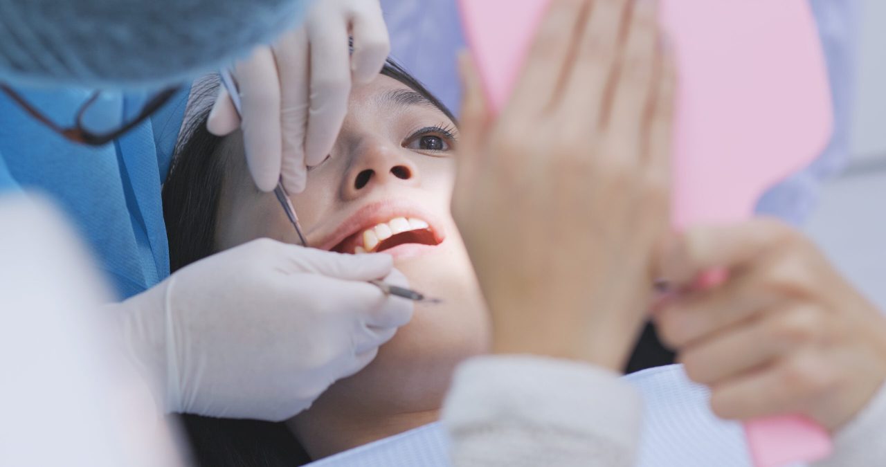Dentista está mostrando para paciente algo nos dentes de trás, ela observa pelo espelho rosa que está segurando.