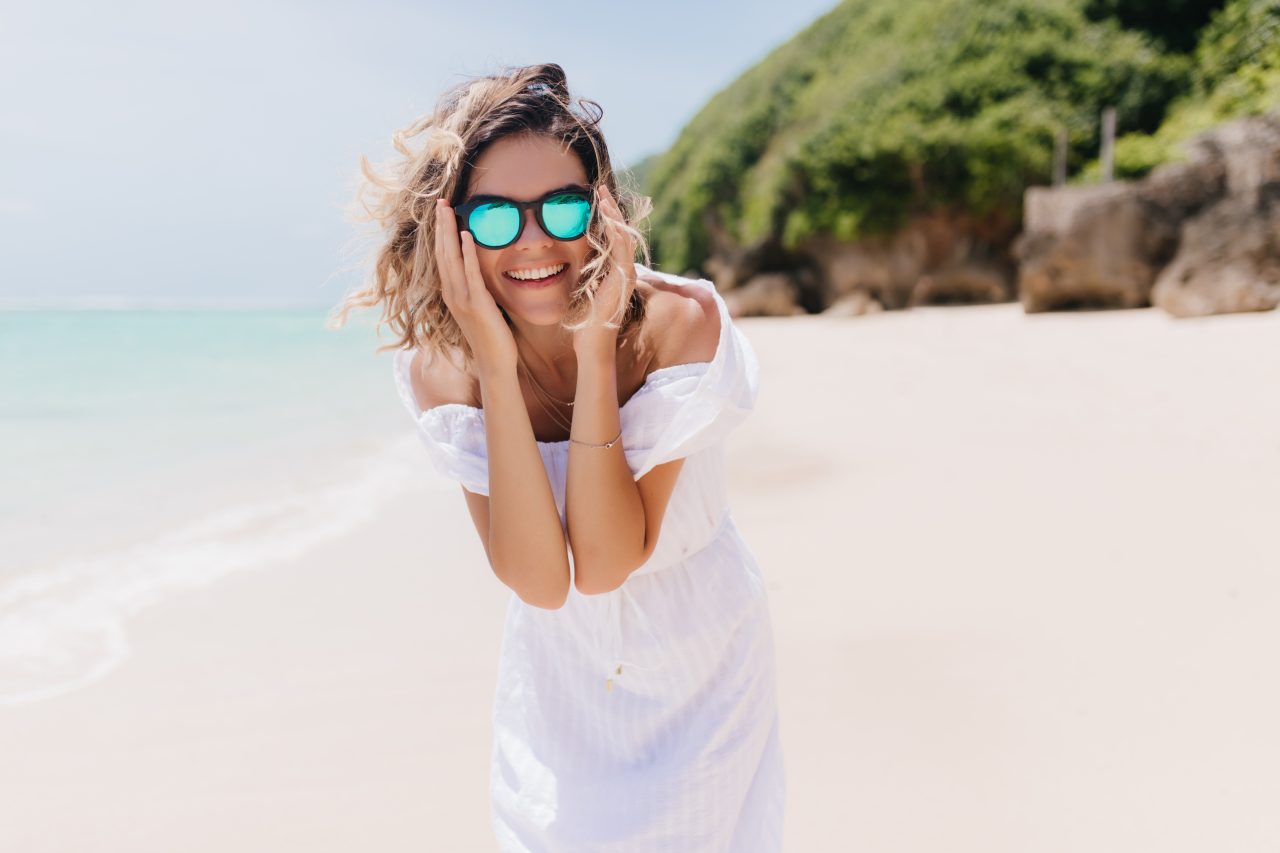 mulher loira usando óculo de sol e vestido branco na praia sorrindo