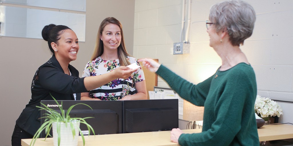 Paciente idosa em clínica odontológica pega papel que secretária sorridente a está entregando.
