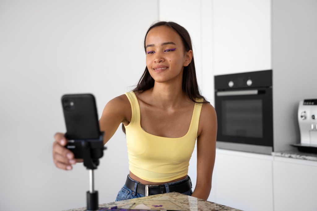 Garota jovem está na cozinha da sua casa colocando um celular no tripé para se gravar falando.