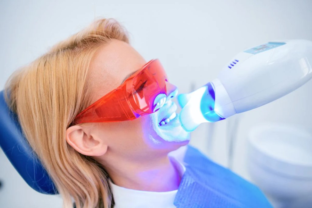 Mulher loira no dentista deitada em cadeira odontológica recebendo tratamento de laser azul para clareamento dental, ela usa um óculos vermelho de proteção