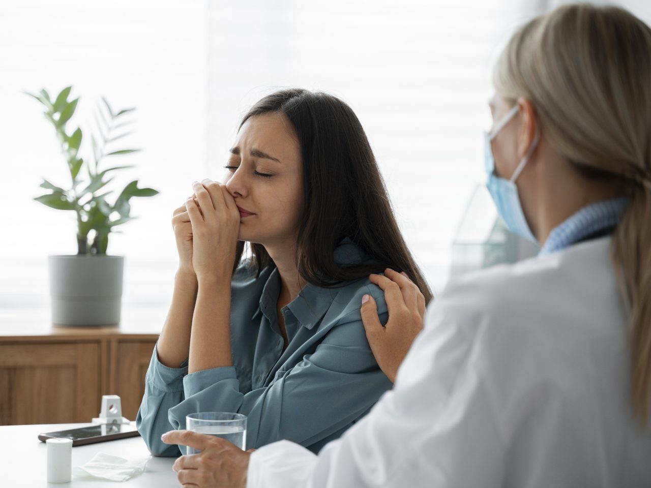 Paciente em consultório chorando, ao lado uma profissional da saúde está oferecendo um copo de água enquanto coloca a mão em seu ombro