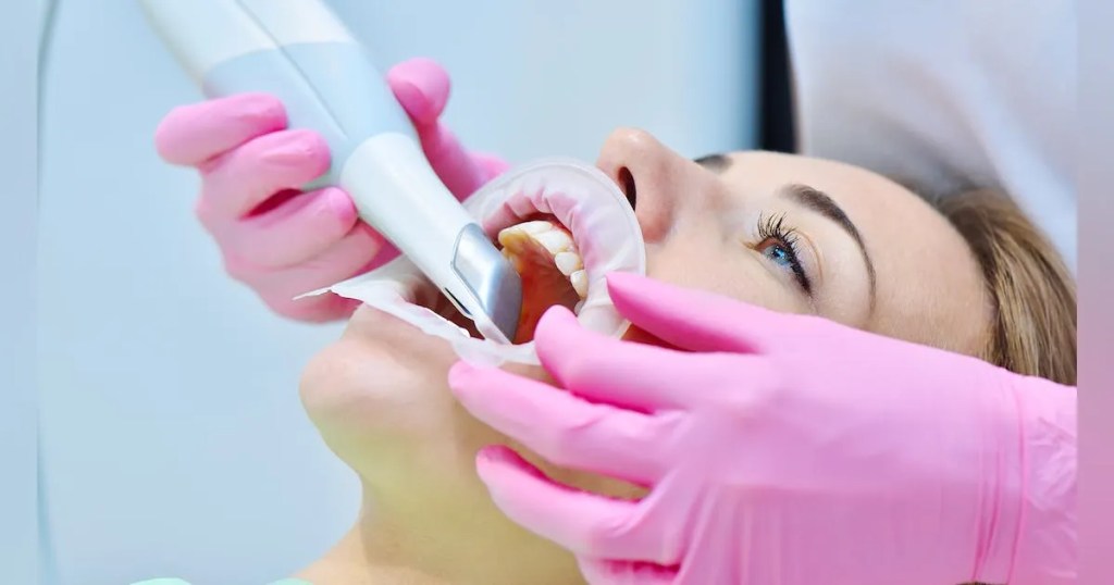 Dentista usando luvas rosas está segurando câmera intaroral realizando escaneamento em boca de paciente.