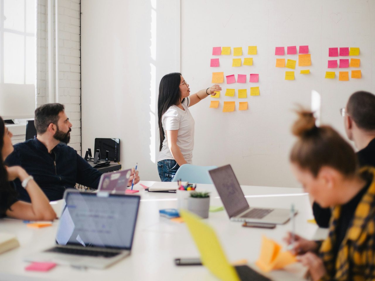 A imagem mostra um sala de reuniões com várias pessoas sentadas a mesa concentradas anotando, com seus notebooks. Na frente tem uma mulher em pé perto do quadro falando e apontando para post-its que estão colados