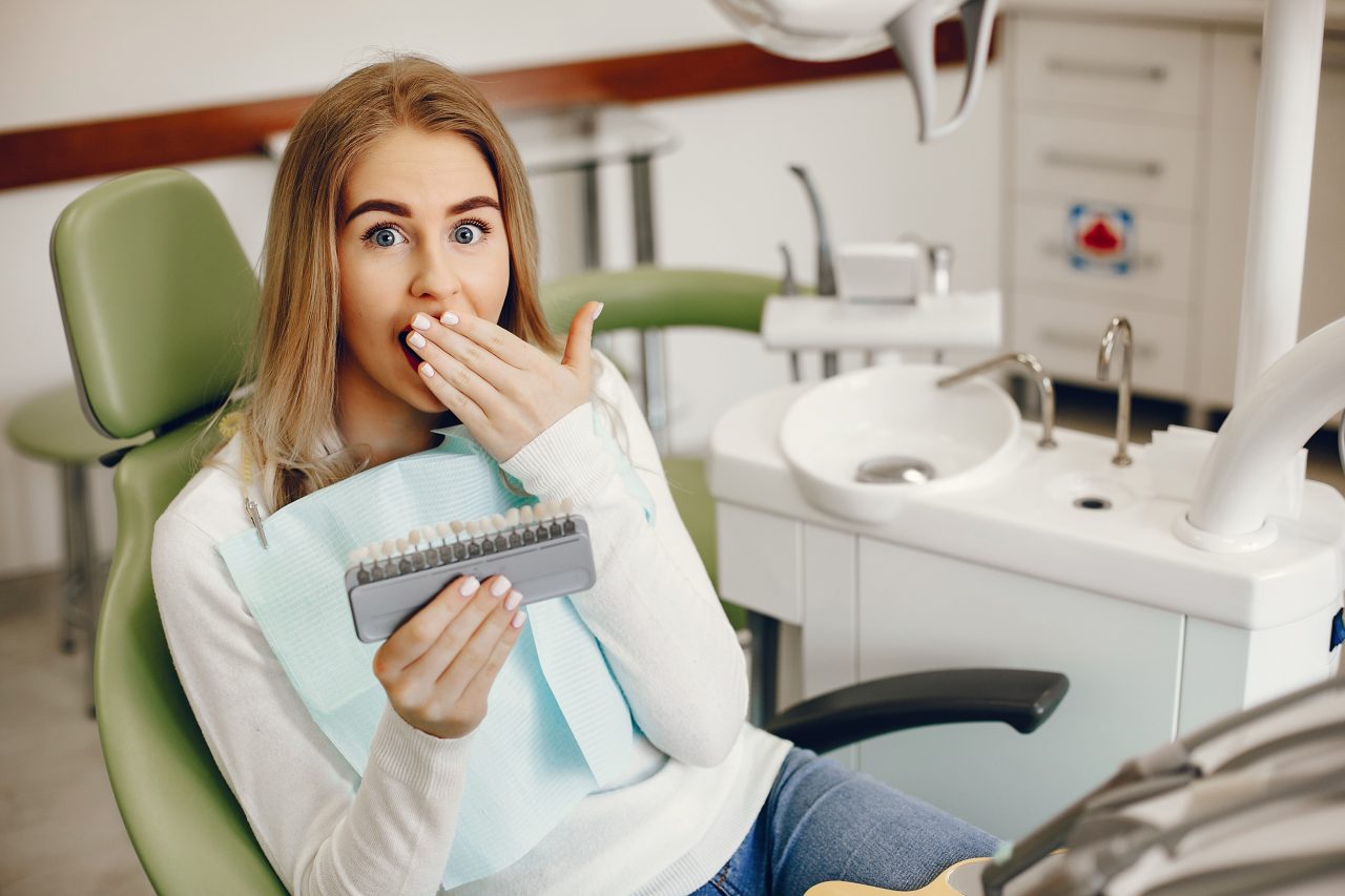 Paciente mulher loira sentada em cadeira odontológica. Ela está com expressão de surpresa e mão em frente a boca, ela está segurando algumas facetas de dente com diferentes cores.