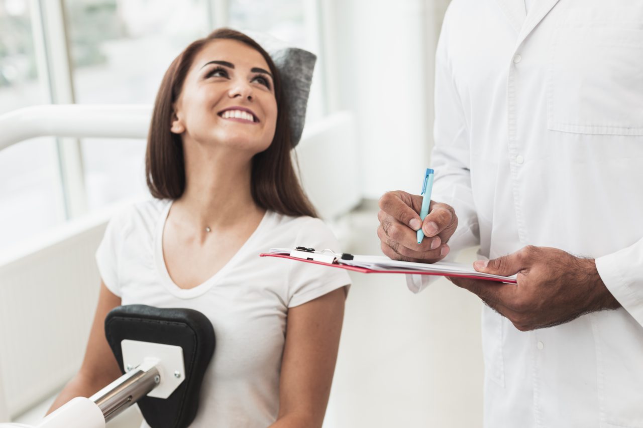 Paciente mulher no dentista, ela está olhando para cima sorrindo. Dentista em pé ao lado dela com prancheta anotando.
