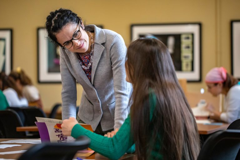 aluna em sala de aula sentada em sua cadeira abrindo o livro e conversando com sua professora que está escorada na mesa
