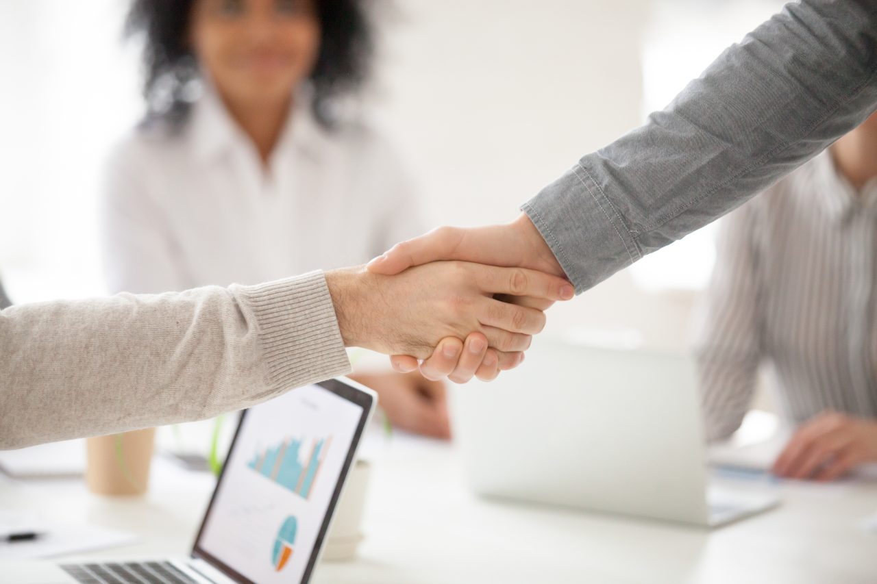 Duas pessoas deram as mãos e se cumprimentaram, no fundo desfocado tem pessoas sentadas em mesa de reunião de escritório