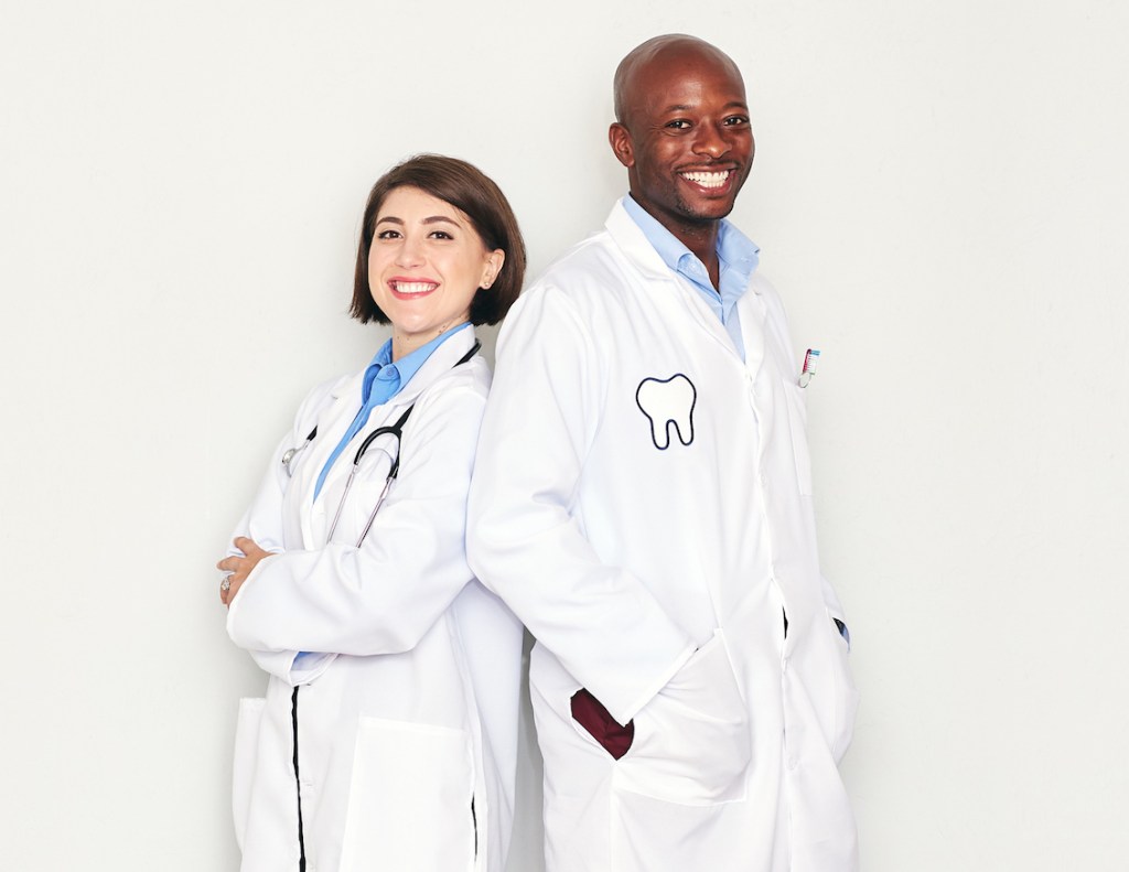 médica com estetoscópio e dentista com jaleco virados costa a costa e sorrindo