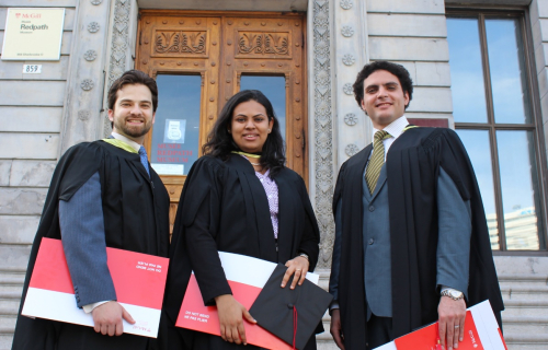 Três alunos com roupas de formaturas e segurando uma pasta com diploma e o capelo