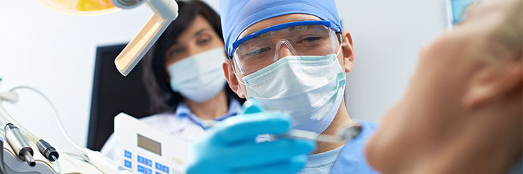 dentista analisando boca de paciente, atrás dele está sua assistente em pé