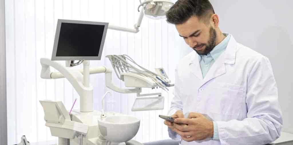 Dentista homem em seu consultório escrevendo uma mensagem de texto no celular enquanto sorri.
