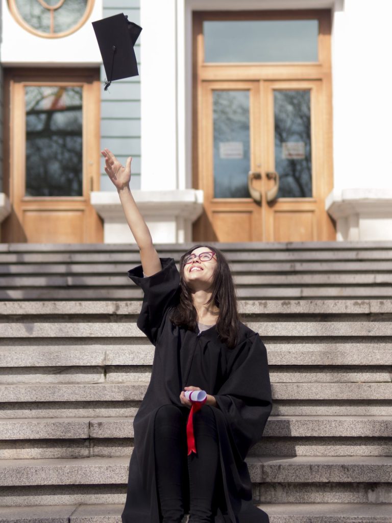Formanda em sua formatura usando beca, segurando o diploma e jogando o capelo para cima