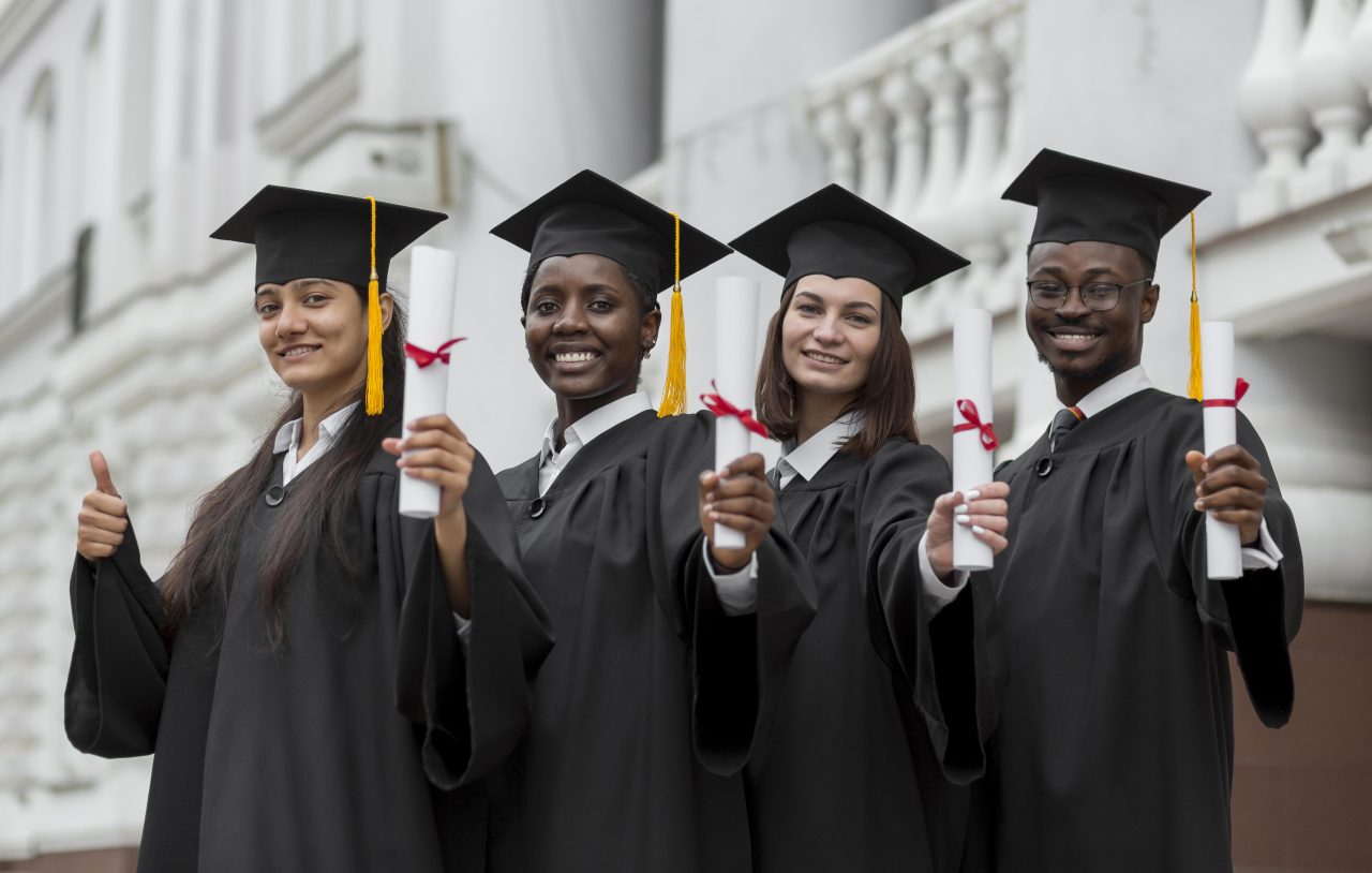 quatro alunos com roupa de formatura segurando seus diplomas e sorrindo