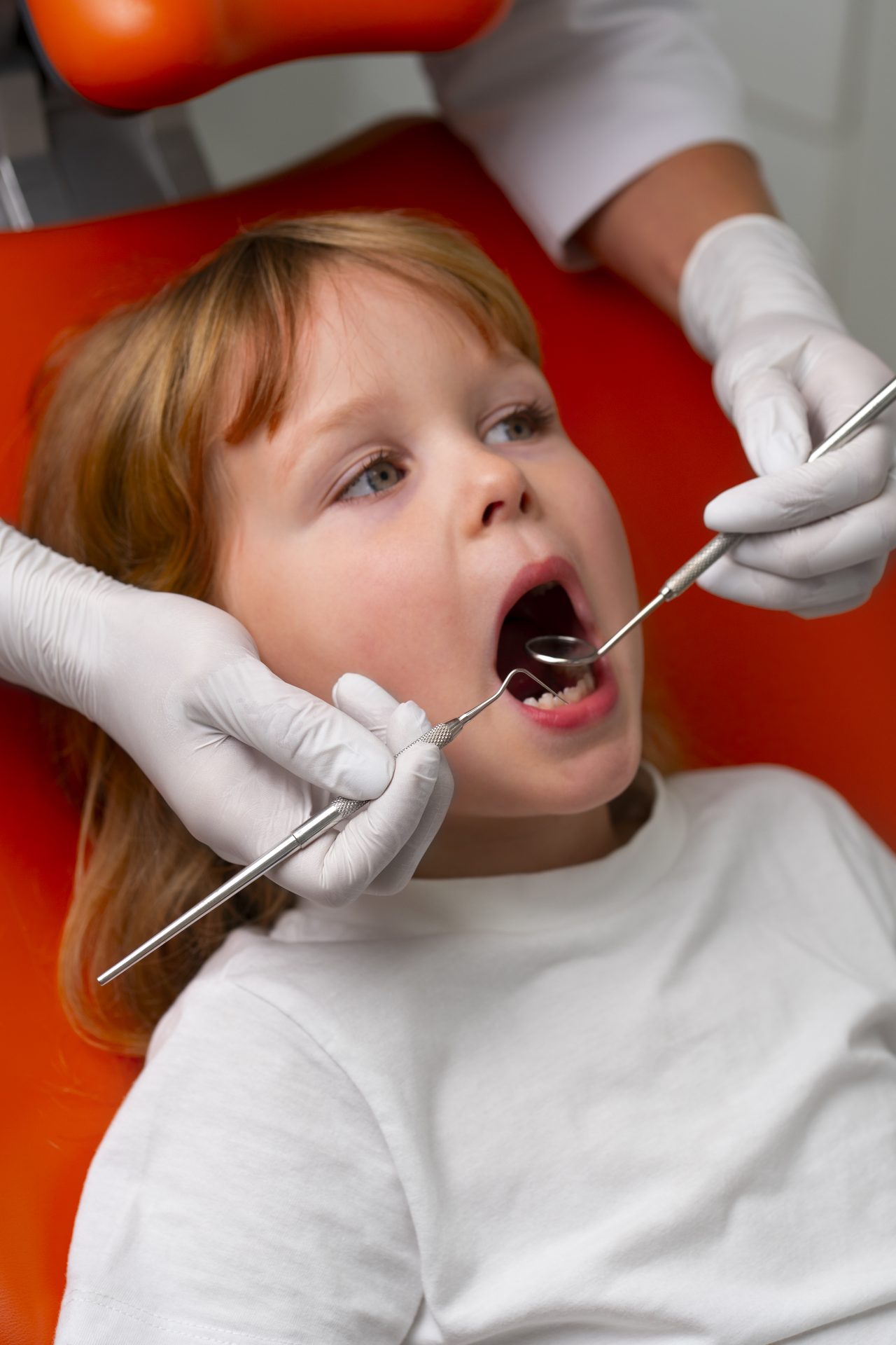 Criança loira em uma cadeira de dentista enquanto o odontopediatra confere se está tudo certo com seus dentes, utilizando um espelhinho