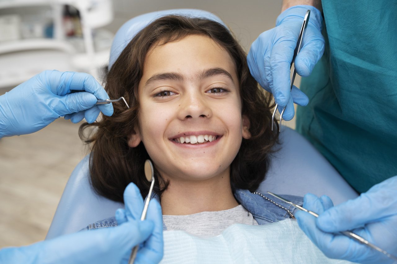 Garoto de cabelo longo sorrindo. Ele está em uma cadeira odontológica, enquanto dois dentistas estão com luvas azuis em volta dele
