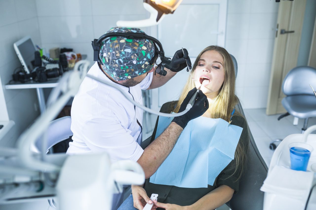Dentista atendendo paciente. Ele usa toca com estampa e luvas pretas. A paciente está deitada na cadeira odontológica.