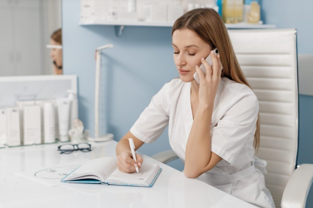 Recepcionista falando em ligação no celular, enquanto anota na agenda. Ela está com uniforme todo branco, tem cabelo liso loiro escuro e no balcão tem cremes.