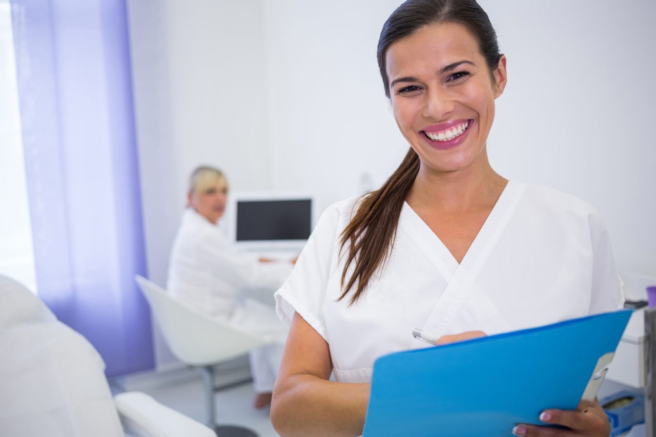 Recepcionista em clínica odontológica sorrindo segurando prancheta azul, no fundo, tem outra recepcionista sentada com mesa com computador