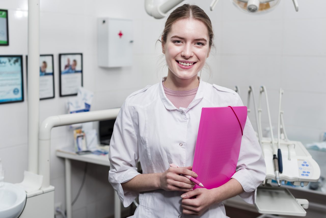 Recepcionista segurando pasta de elástico rosa e caneta e sorrindo para câmera, ela está dentro do consultório