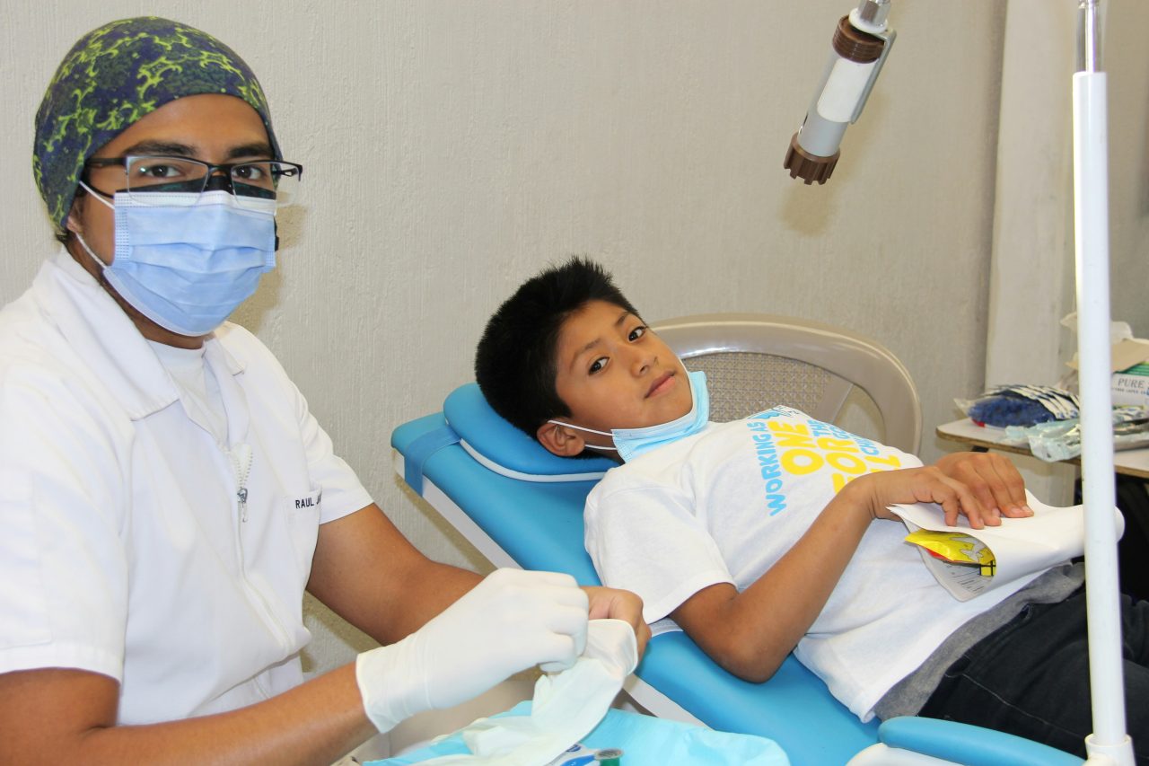 Dentista sentado ao lado de cadeira odontológica com paciente criança.