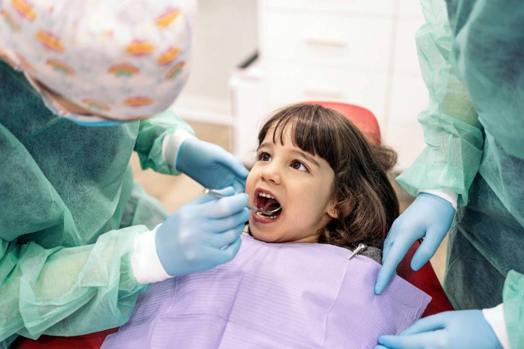 Dentista e assistente examinando dentes de criança com espelhinho.