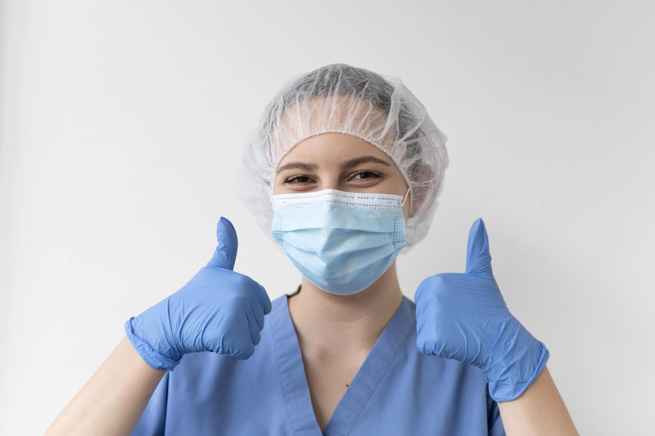 Dentista mulher usando touca descartável, máscara e luva, está feliz e fazendo sinal de positivo com as duas mãos