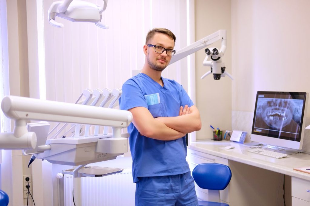 Dentista homem está de pé em seu consultório odontológico, no computador está aberto a imagem de um panorâmica