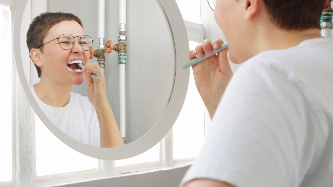 Mulher de cabelo curtinho, blusa branca e óculos de grau, está escovando os dentes