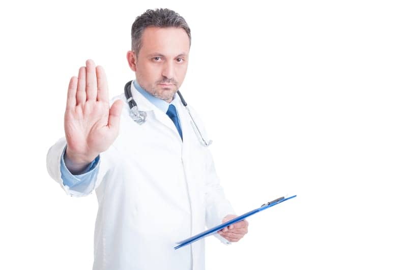 Médico de expressão séria, segura uma prancheta com uma mão e a outra ele está com a palma levantada em sinal de pare.