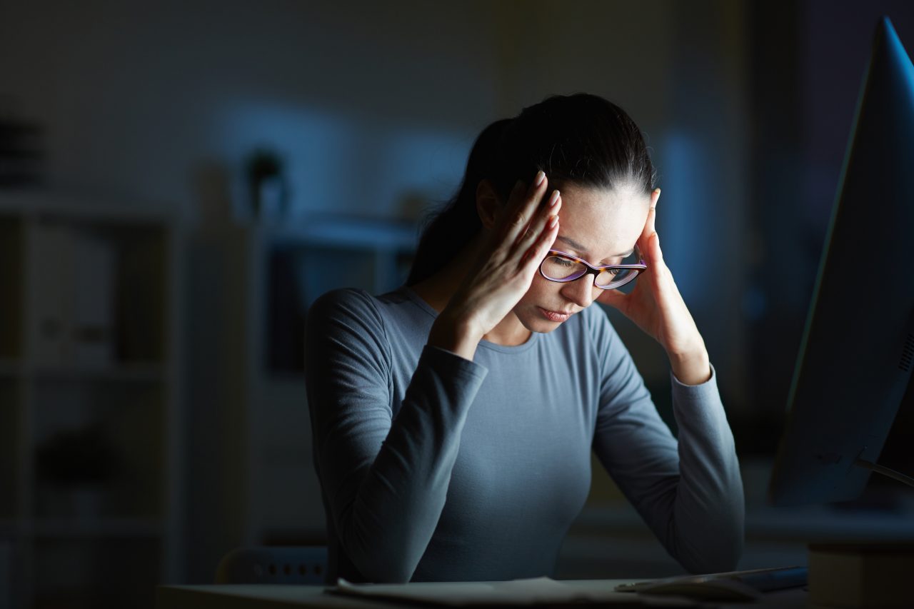 Mulher em quarto escuro, com apenas a luz do computador em seu rosto. Ela está com as mãos na testa e olhos fechados, demonstrando exaustão. Imagem pra demonstrar o sentimento da síndrome de burnout.